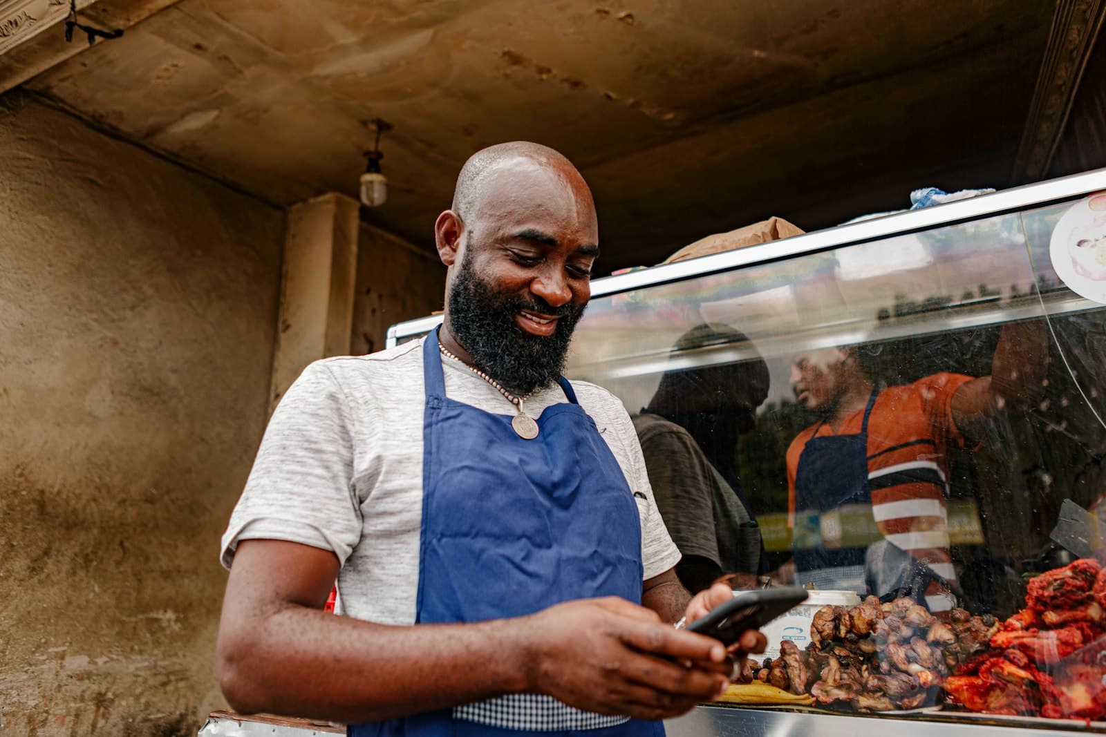 Shop owner in an apron smiling at his phone behind a food counter